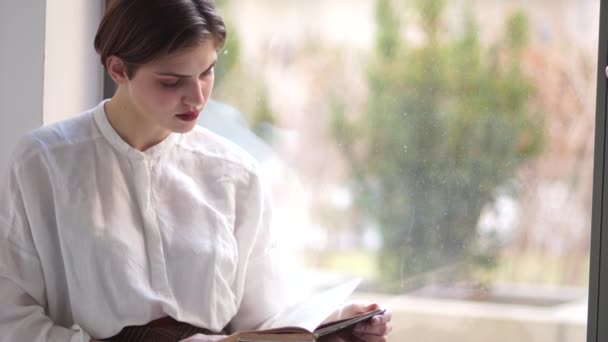 Un retrato cercano de una chica con un corte de pelo corto es leer un libro viejo mientras está sentada en un alféizar de la ventana. Mujer feliz — Vídeos de Stock