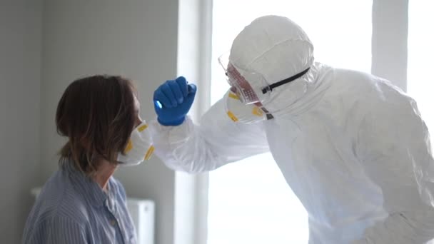 Dokter met een beschermend masker en bril onderzoekt een vrouw met een zaklamp. Pandemische dreiging, coronasirus bedreiging — Stockvideo