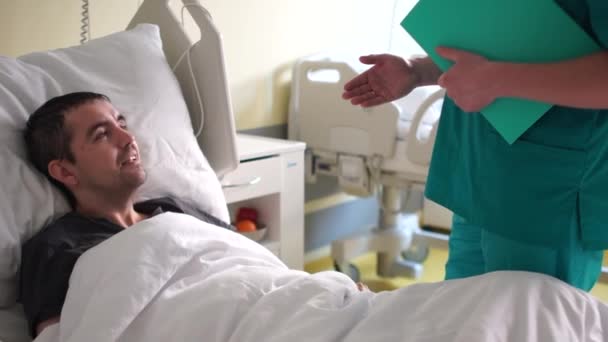 Реабилитация после инсульта. Молодой человек лежит в постели, рядом с ним стоит врач, пациент разговаривает с врачом — стоковое видео