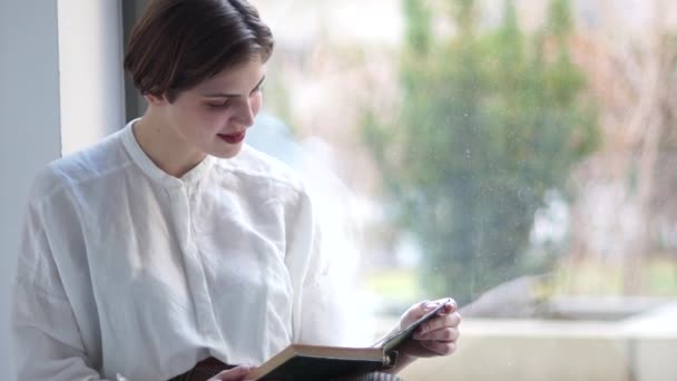 Close Indore portrait of a young girl in a vintage white blouse. A woman is sitting at the window and reading a book. Happy woman, calm weekend, meditation and self-knowledge. — Αρχείο Βίντεο