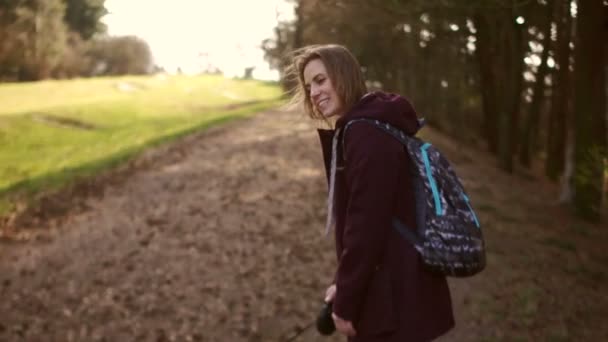 Egy fiatal nő pórázon sétáltatja a kutyáját egy tavaszi városi parkban. A lány burgundi dzsekiben van, és hátizsákot visel a hátán. — Stock videók