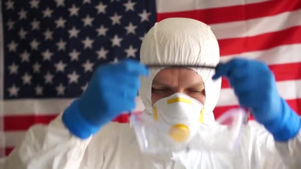 O jovem em um terno protetor põe uma máscara contra o fundo da bandeira do estado dos EUA. Epidemia de coronovírus nos EUA, covid-19. Prevenção de epidemias — Vídeo de Stock