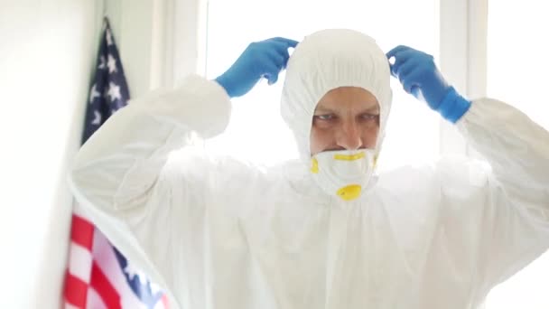 Propagación del coronavirus en los Estados Unidos. Prevención de epidemias y control de virus. Un hombre con un traje protector se pone una máscara, junto a la bandera del estado de EE.UU. — Vídeo de stock