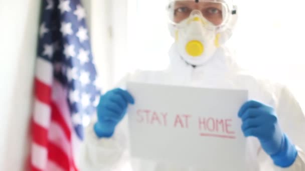 Médico estadounidense con traje protector y máscara en el fondo de la bandera del estado de Estados Unidos. Un hombre sostiene una foto con la inscripción en casa. Coronovirus covid19 y cuarentena en América — Vídeo de stock