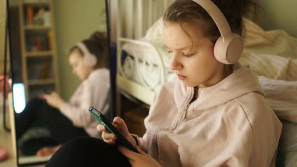 Egy tinédzser lány okostelefonnal a kezében és vezeték nélküli fejhallgatóval zenét hallgat. Karantén hobbik, gyermekek és szerkentyűk — Stock videók