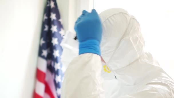Koruyucu giysili Amerikalı doktor, ABD bayrağının arka planına karşı bir maske takıyor. Yan görüş. Coronovirus Amerika 'da 19 yaşında. Virüsün yayılmasını engellemek ve karantinaya almak. — Stok video