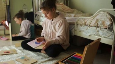 Güzel kız odasında yerde otururken pastel boyalarla resim çizer. Karantina ve kendini izole etme süresince mutlu bir genç. Çocukların yaratıcılığı