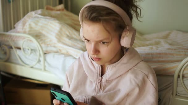 Urocza, kręcona nastolatka ze smartfonem w rękach i bezprzewodowymi słuchawkami słucha muzyki. Zainteresowania kwarantanną, dzieci i gadżety. Zamknij Indore portret — Wideo stockowe