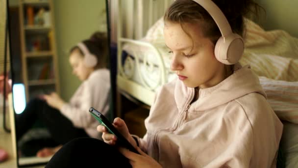 Chica adolescente con un teléfono inteligente en sus manos y auriculares inalámbricos escuchando música. Pasatiempos de cuarentena, niños y gadgets — Vídeos de Stock