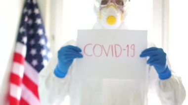 Adamın elinde kırmızı renkli Covid-19 yazan bir tabela var. Birleşik Devletler bayrağının arka planında tıbbi koruyucu bir antiviral giysi giymiş bir adam. ABD 'de koronovirüsle mücadele.