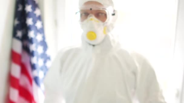 男は碑文と写真を保持している家に滞在します。米国国旗を背景に防護服とマスクを着たアメリカ人医師。コロノウイルスのコビド19とアメリカの隔離 — ストック動画