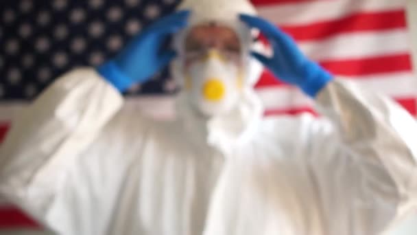 Clouse up mladý muž v ochranném obleku nasadí masku na pozadí amerického státu vlajky. Koronovirová epidemie v USA, covid-19. Prevence epidemie — Stock video