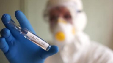 Koruyucu kıyafetli, maskeli ve gözlüklü bir mikrobiyolog elinde aşı olan bir ampul tutuyor. Coronovirus covid-19 aşısı arayın. Coronovirüs aşısı