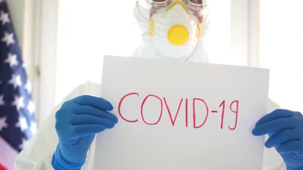 米国でのCovid19コロナウイルス流行。Covid19と書かれたサインを持っている保護スーツの男の近しい肖像画。隔離と隔離 — ストック動画