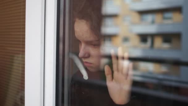 КОВИД 19 Изоляция. Оставайся дома на карантине. Красивая девочка-подросток смотрит в окно на улицу. Грустный ребенок дома во время самоизоляции — стоковое видео