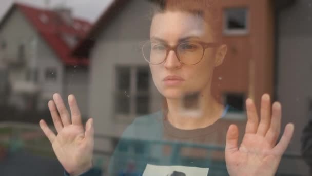 Muchacha triste con gafas mira por la ventana a la calle. Cuarentena durante la epidemia de coronavirus covid-19 — Vídeo de stock