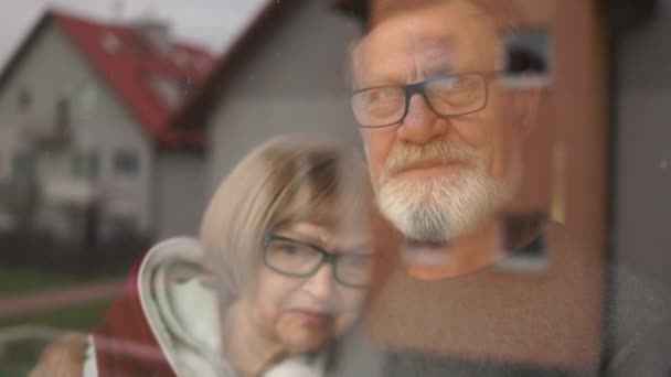 Starsza rodzina emerytów, mąż i żona, stoją przy oknie i patrzą na ulicę, uśmiechając się smutno. Izolacja podczas kwarantanny koronowirusa covid-19 — Wideo stockowe