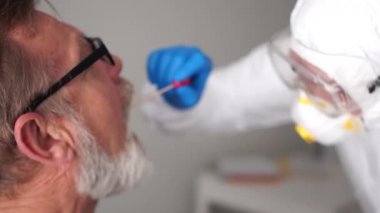 Koruyucu takım elbiseli bir doktorun portresi yaşlı bir emeklinin boğazından örnek alıyor. PCR reaksiyonundan zararsız tanı yöntemi