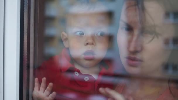 Genç aile, anne ve iki çocuk pencerede duruyorlar. Covid-19 koronavirüs salgını sırasında karantina ve kendini izole etme — Stok video