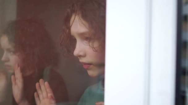 Gyerekek a karantén alatt. Két szomorú göndör gyerek, testvérek, egy fiú és egy lány egy pohár mögött az ablak mellett. Karantén Coronovírus Covid-19 — Stock videók