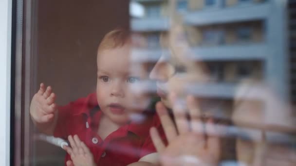 Concetto di quarantena e autoisolamento. Il bambino tra le braccia di sua madre sta toccando il vetro. Famiglia vicino alla finestra durante la quarantena — Video Stock