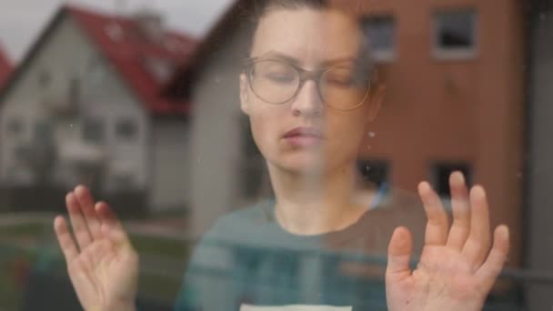 Νεαρή θλιμμένη γυναίκα κοιτάζει έξω από το παράθυρο. Αυτοαπομόνωση και καραντίνα από τον ιό Covid-19. Κλείσιμο πορτραίτου μιας γυναίκας με γυαλιά πίσω από ένα παράθυρο — Αρχείο Βίντεο