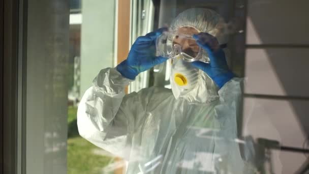Koruyucu kıyafetli çok yorgun bir doktor maskeyi çıkarır ve eliyle cam pencereye yaslanır. Doktorlar sıkı çalışma konsepti. Salgın koronovirüs covid-19, tıbbi çöküntü. — Stok video
