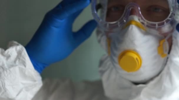 Microbioloog in een beschermend pak doet een masker op en neemt een buis met een monster van bloedplasma in zijn hand. Laboratoriumstudies van het vaccin tegen het coronovirus Covid-19 — Stockvideo