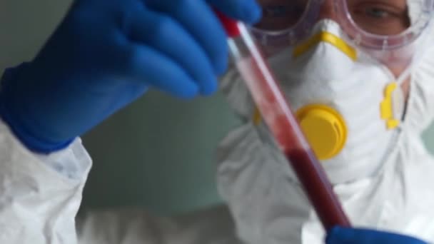El microbiólogo masculino con traje protector y máscara tiene en la mano un tubo de ensayo con una muestra de plasma sanguíneo. Estudios de laboratorio de una vacuna contra el coronovirus — Vídeo de stock