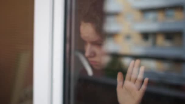 Αυτοαπομόνωση έννοια. Μια θλιμμένη μαθήτρια στέκεται στο παράθυρο και κοιτάζει το δρόμο, χτυπώντας το γυαλί με το χέρι της. Κατάθλιψη κατά την καραντίνα coviro- 19 — Αρχείο Βίντεο