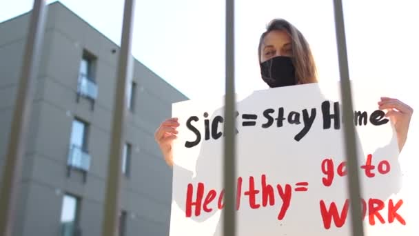 Fille dans un masque noir avec une affiche malade rester à la maison, aller en bonne santé au travail. La manifestation pendant la quarantaine du coronovirus près de la clôture des institutions de l'État. Restriction de quarantaine grève — Video