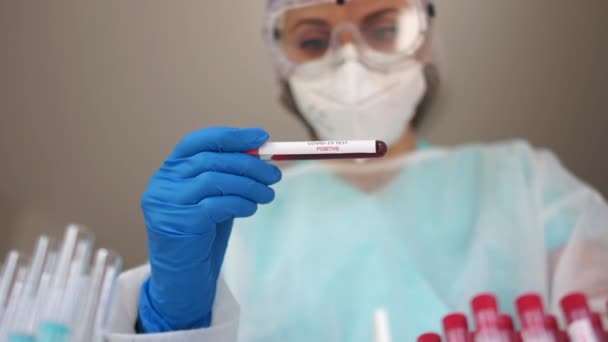 Микробиолог в лаборатории держит пробирку с образцом крови, инфицированным короновирусным ковидом-19 сарс-ков-2. Надпись на пробирке ковид-19 положительная. Положительный короновирусный тест — стоковое видео