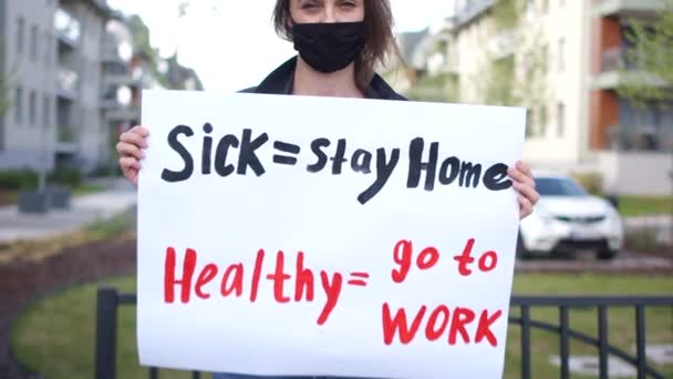 Ragazza attivista con un poster malato - rimanere a casa, sano - andare a lavorare in mano. La donna indossa una maschera protettiva nera, protesta durante la quarantena del coronovirus covid-19 — Video Stock