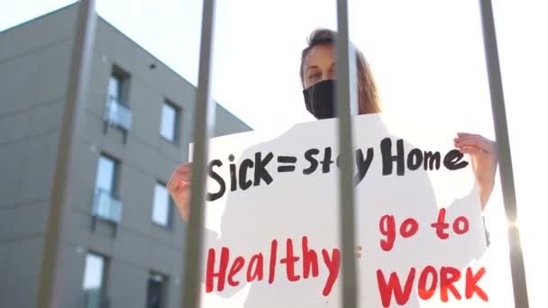 Больной - оставайтесь дома здоровым - идти на работу. Солнечный портрет молодого студента-активиста в черной маске с плакатом против продолжения карантина-19 — стоковое видео