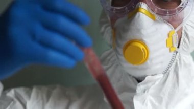 Koruyucu kıyafetli ve elinde deney tüpü olan bir maske takmış erkek bir mikrobiyolog portresi. Kan plazma örneği, Covid-19 koronovirüs testi.