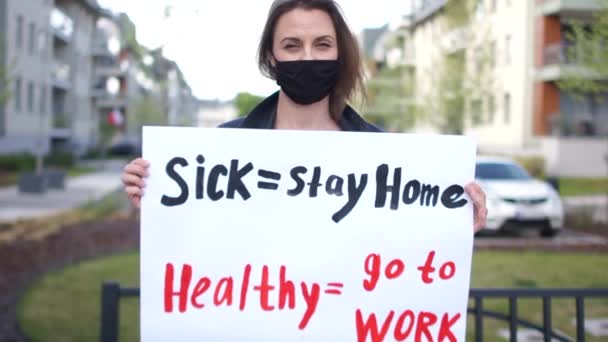 Больной - оставайтесь дома здоровым - идти на работу. Девушка-активистка с плакатом в руках. Женщина носит черную защитную маску, протестует во время карантина короновируса ковид-19 — стоковое видео
