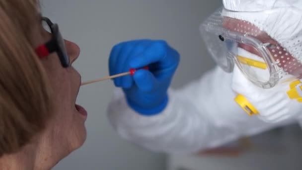 Coronovirus-Test, Test zur Bestimmung einer Coronovirus-Infektion. Mikrobiologe in einem Schutzanzug entnimmt eine Probe von Biomaterial aus der Mundhöhle einer älteren Frau — Stockvideo