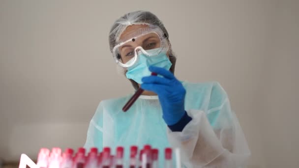 Κορίτσι βοηθός εργαστηρίου σε μια προστατευτική στολή, μάσκα και γάντια κρατά ένα δοκιμαστικό σωλήνα με ένα δείγμα αίματος μολυσμένο με covid Covid-19. Διαγνωστικά του sars-cov-2 — Αρχείο Βίντεο