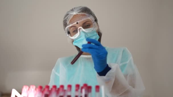 Menina assistente de laboratório em um terno de proteção, máscara e luvas detém um tubo de teste com uma amostra de sangue infectado com coronovírus Covid-19. Diagnóstico de sars-cov-2 — Vídeo de Stock