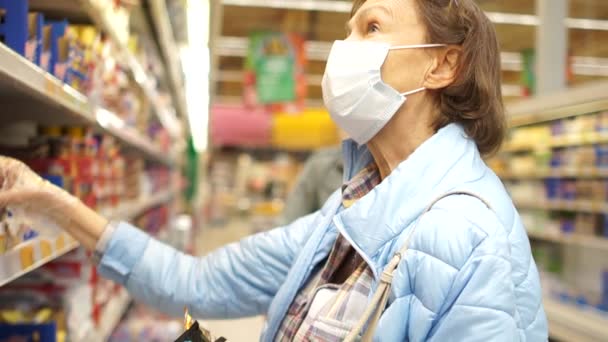 一位戴着面具和手套的老年妇女选择超市货架上的产品。在Coronovirus covid-19的检疫过程中，她正在购买一位孤独的家庭主妇。社会距离 — 图库视频影像