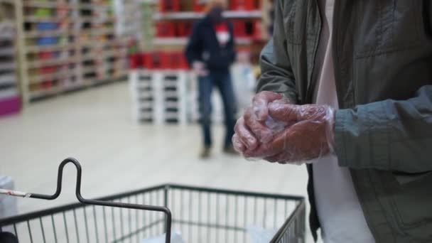 Šedovousý důchodce s brýlemi v masce a rukavicemi nese vozík mezi regály velkého obchodu. Pravidla pro návštěvu supermarketů po skončení karantény — Stock video