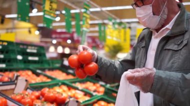 Cerrahi maskeli ve plastik eldivenli olgun bir adam, Coronavirus covid-19 karantinası sırasında süpermarketten alışveriş yapıyor. Yaşlı vatandaş süpermarketten domates topluyor ve onları el arabasına koyuyor.