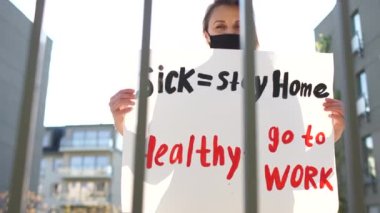Hasta - evde sağlıklı kal - işe git. Avrupa, Coronavirus covid-19 karantinası sırasında katı tecrit tedbirlerine karşı protestolar düzenliyor. Belediyenin çitlerinin yanında posterli maskeli bir kız duruyor.
