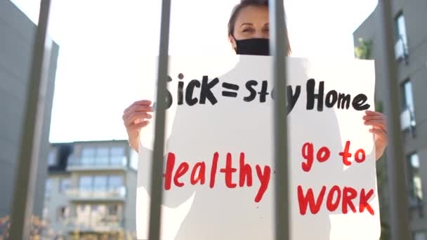 生病了- -健康地呆在家里- -去工作.欧洲抗议在检疫Coronavirus covid-19期间采取严格的封锁措施。戴着面具、贴着海报的女孩站在市政厅栅栏旁边 — 图库视频影像