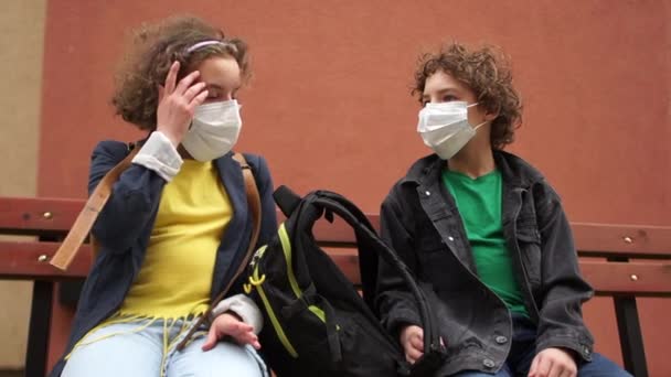 Torniamo a scuola dopo la quarantena. carino riccio ragazzo e ragazza sono seduti su un panchina a una distanza di sicurezza e parlando — Video Stock