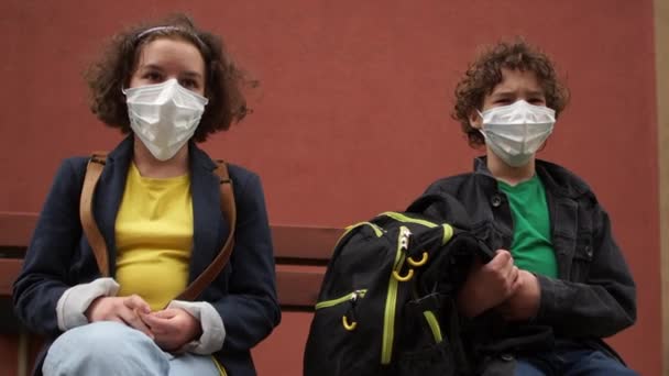 隔離後学校に戻る。かわいい巻きマスクの男の子と女の子は安全な距離でベンチに座って話しています。検疫後の生活 — ストック動画