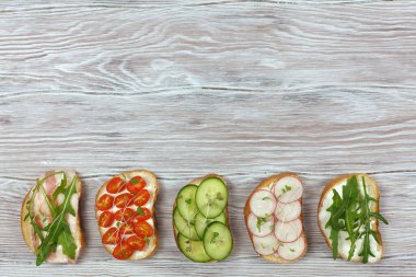 Krem peynirli, sebzeli, pastırmalı ve mikroyeşillikli mini sandviç koleksiyonu. Peynirli sandviçler, salatalık, turp, domates, domuz pastırması, roka ve açık ahşap zemin üzerinde mikroyeşil. Üst Manzara. Düz yatıyordu.