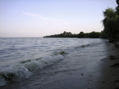 Kumlu sahilde soluk bir dalga kırılıyor. Ağaçlar ve çalılar kıyıda yetişir. Geç oluyor..