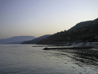 Sakin bir denizin kıyısındaki dalgalı su... kıyı dağlarının arka planında... gökyüzünün sisinde... akşam vakti....