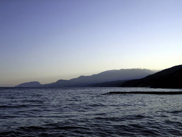 夕方の距離で空のもやの中で沿岸の山々の背景に穏やかな海の岸に防波堤 — ストック写真